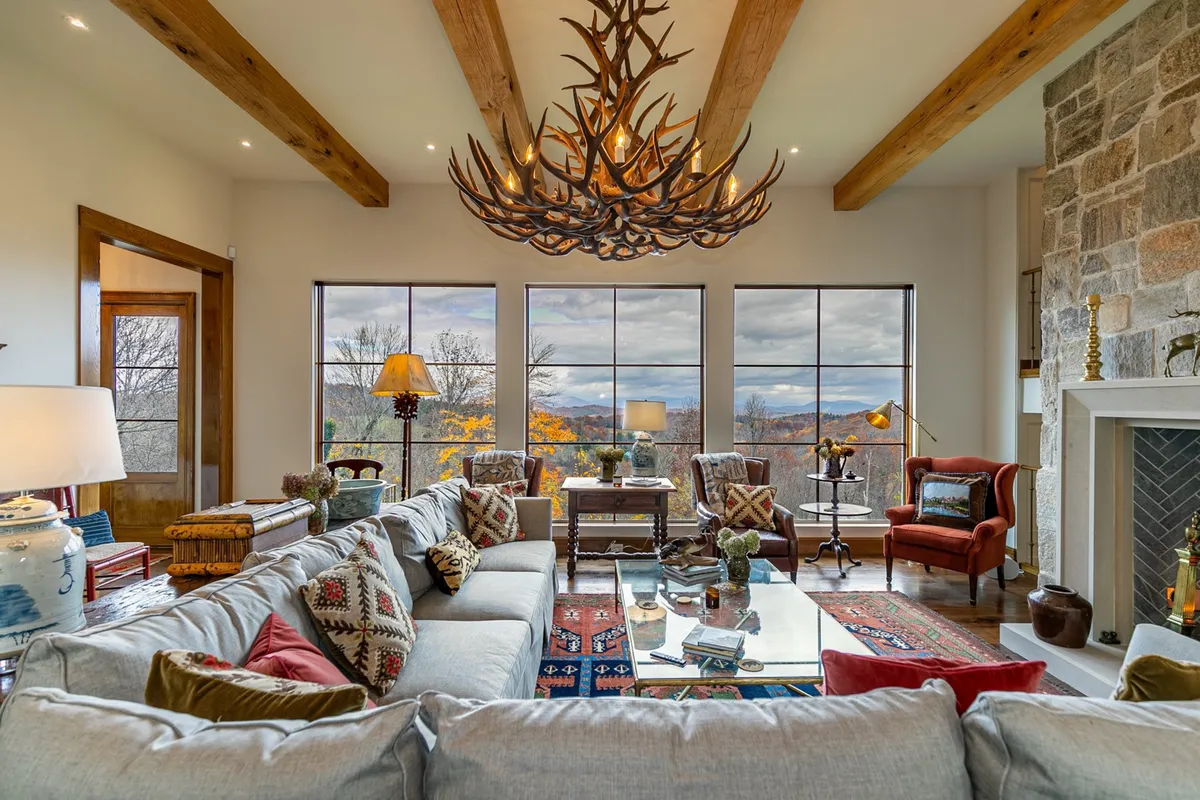 This image features a large antler chandelier illuminating a spacious great room with a comfortable sectional sofa, stylish armchairs, and a decorative coffee table. Expansive windows provide a panoramic view of the surrounding mountains, inviting natural light and scenic beauty into the heart of the lodge.