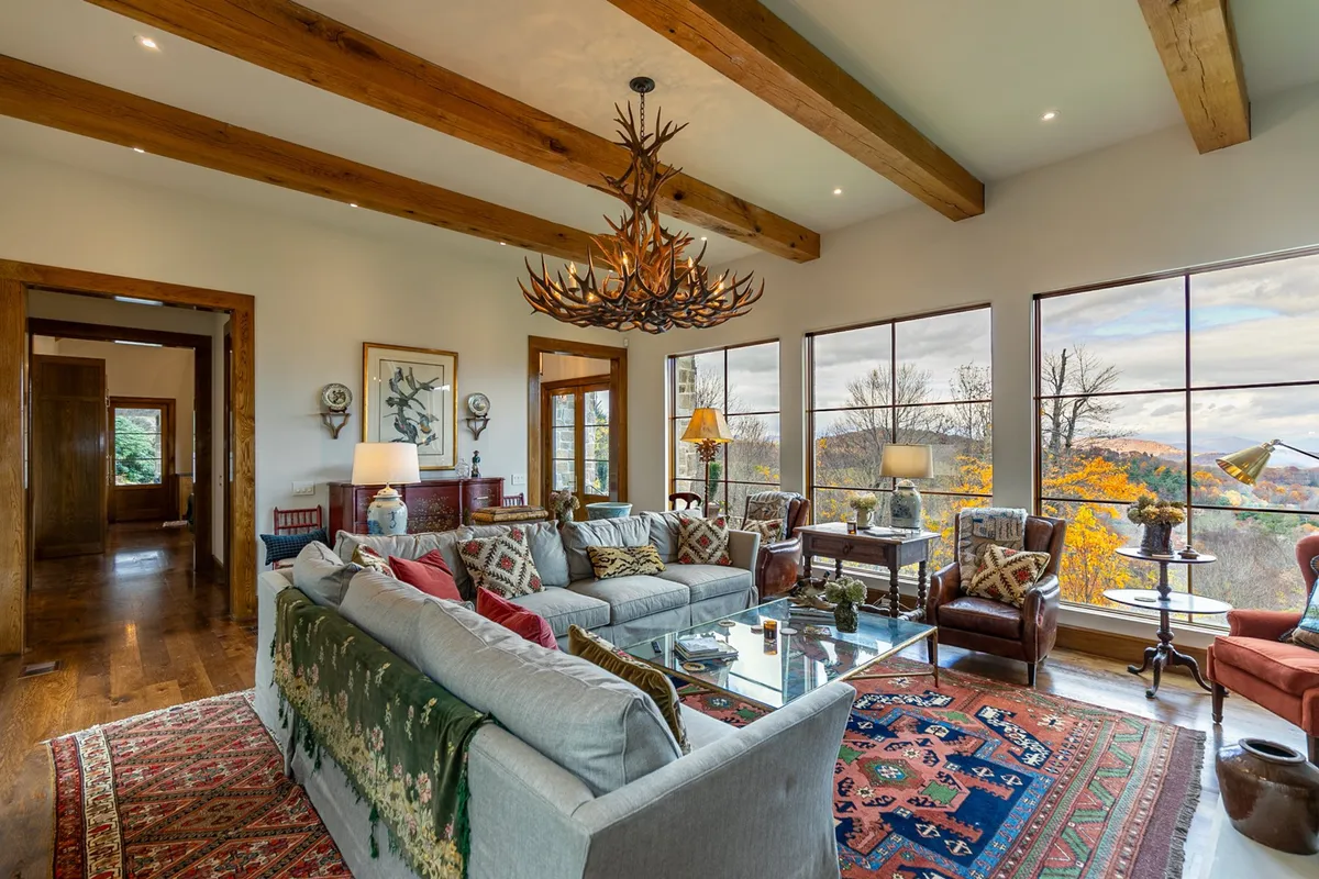 A spacious living area features a large, comfortable sofa with patterned cushions, an eye-catching antler chandelier, expansive windows with scenic views, a decorative rug, and stylish armchairs arranged for group gatherings. The ambiance is cozy and perfect for hosting family or friends.