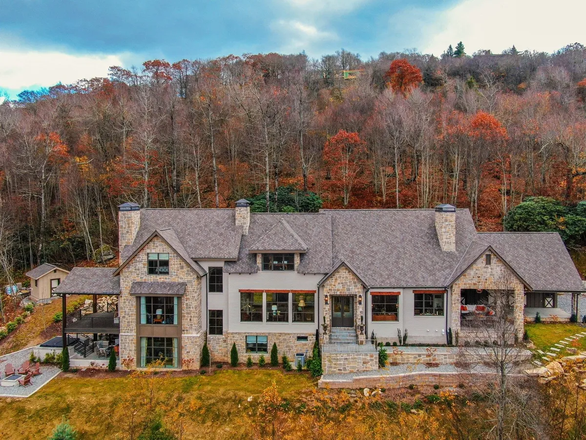 A wide exterior view of the home’s stone and wood architecture, expansive patio, and large windows, all surrounded by vibrant autumn foliage. The setting showcases areas for outdoor dining, family gatherings, and quiet moments embraced by nature.