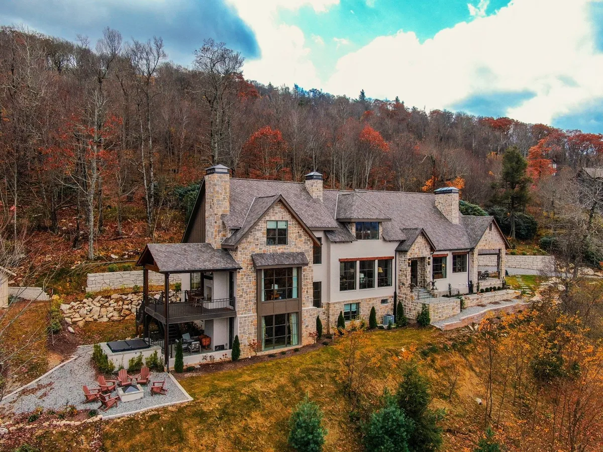 The image showcases a grand, custom-built stone-clad house nestled among tall trees, with a spacious outdoor seating area perfect for gatherings. The surrounding forest provides a tranquil backdrop, while cozy seating and a stone fire pit invite guests to enjoy evenings outdoors. Sunlight filters through the trees, illuminating the inviting patio and hinting at nearby walking trails that beckon exploration.