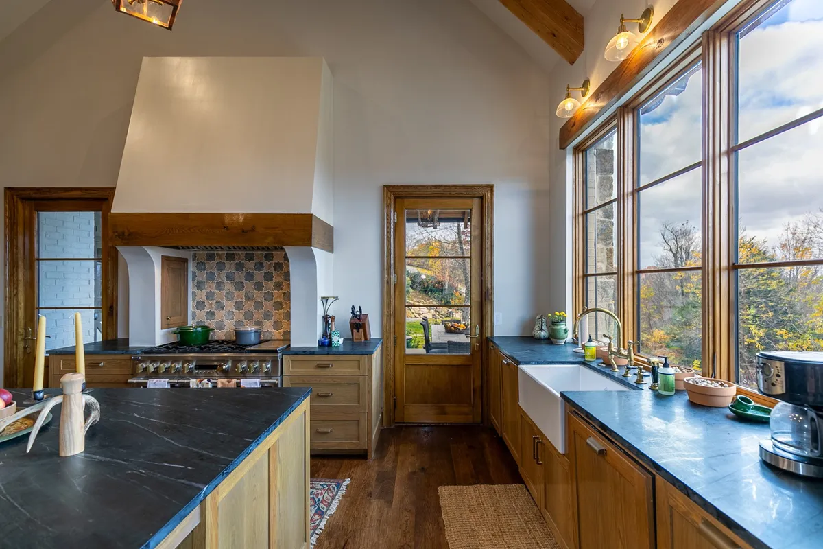 A view of the gourmet kitchen, featuring a spacious island, premium stove with hood, deep sink, and large windows that invite abundant natural light. The kitchen’s design combines luxury with warmth, showcasing soapstone countertops and wood cabinetry, ideal for both culinary adventures and socializing.