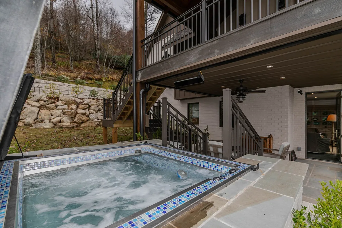 An inviting exterior space featuring a hot tub set into a landscaped area, with stone walls, an outdoor seating area, and steps leading to an upper deck. The scene captures a tranquil and social mood, ideal for relaxing in the hot tub while enjoying mountain sunsets or entertaining guests outdoors.