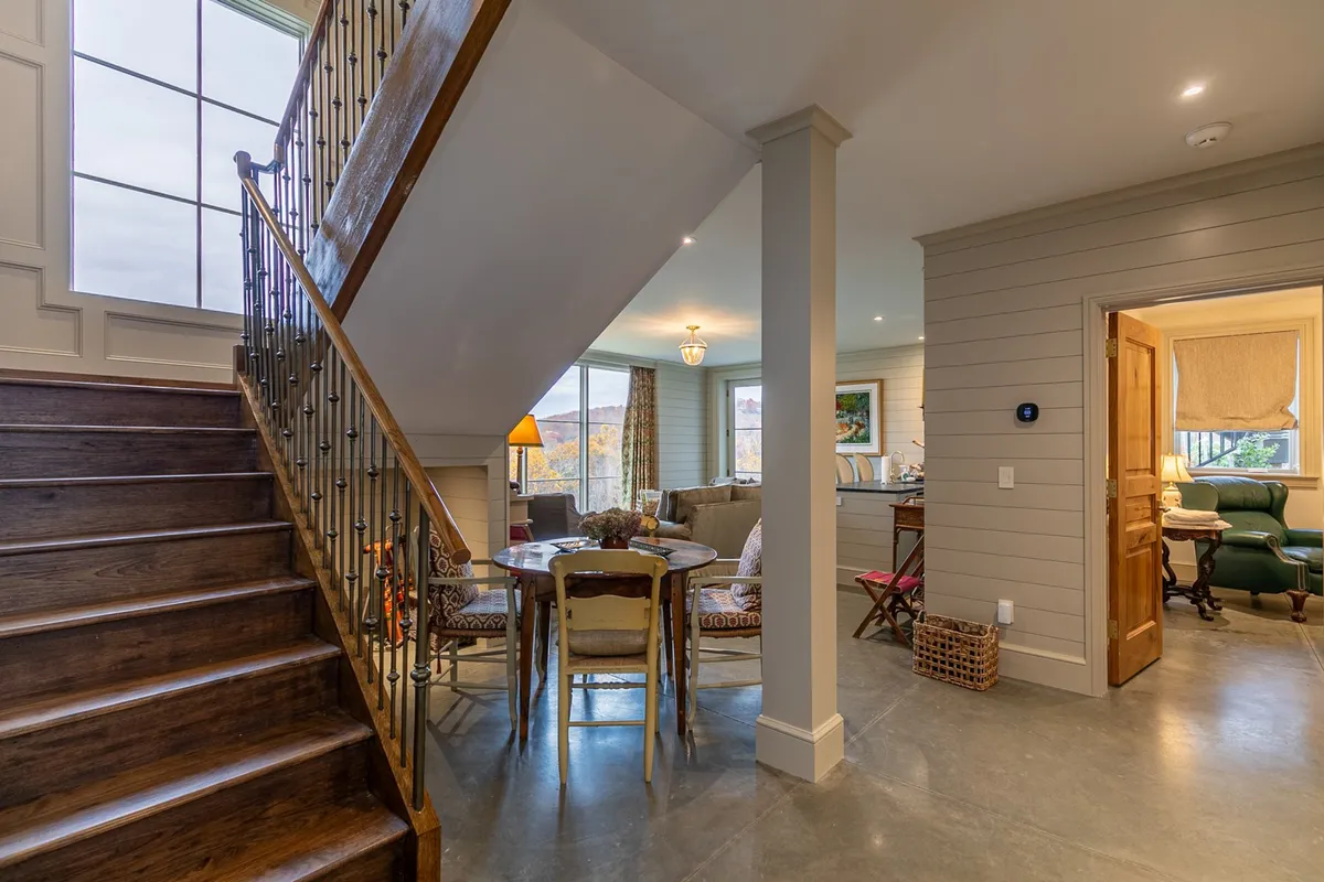 An open-plan space showcasing a dining area with a large table, a cozy seating area, and a staircase leading to other parts of the lodge. A large window offers views and natural light, reinforcing a sense of spaciousness and connection.