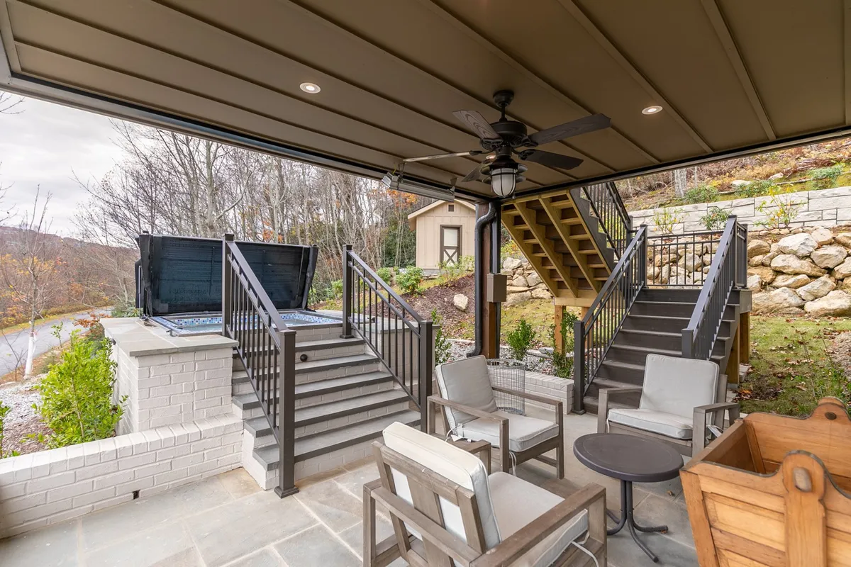 This exterior shot features a comfortable patio seating area with a hot tub, a staircase leading to the upper level, and lush greenery all around. The setting is perfect for entertaining, soaking in panoramic mountain views, or gathering around the fire pit at dusk.