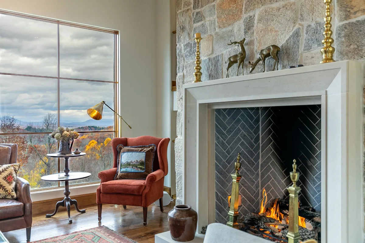 A cozy seating area with two armchairs positioned in front of a stone fireplace, set beside a large window that offers a sweeping view of the outdoors. The space is filled with natural light, creating an inviting environment for relaxing and watching the changing seasons.