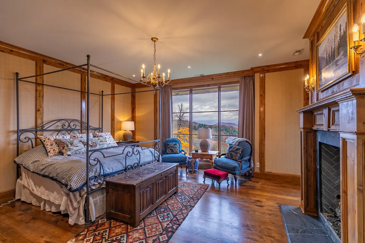 A beautifully appointed bedroom featuring a four-poster bed, a cozy fireplace, large windows offering mountain views, and comfortable blue armchairs. The décor is serene and elegant, creating a sense of a private retreat.