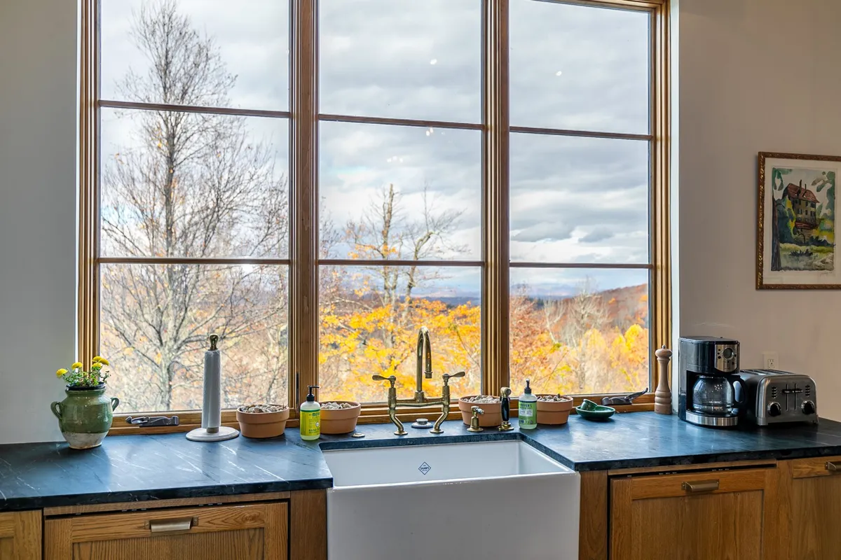Large windows frame a scenic view, with a farmhouse sink and decorative items on the countertop. The kitchen is oriented toward the outdoors, inviting guests to admire nature while preparing meals and socialize in a bright, airy setting.