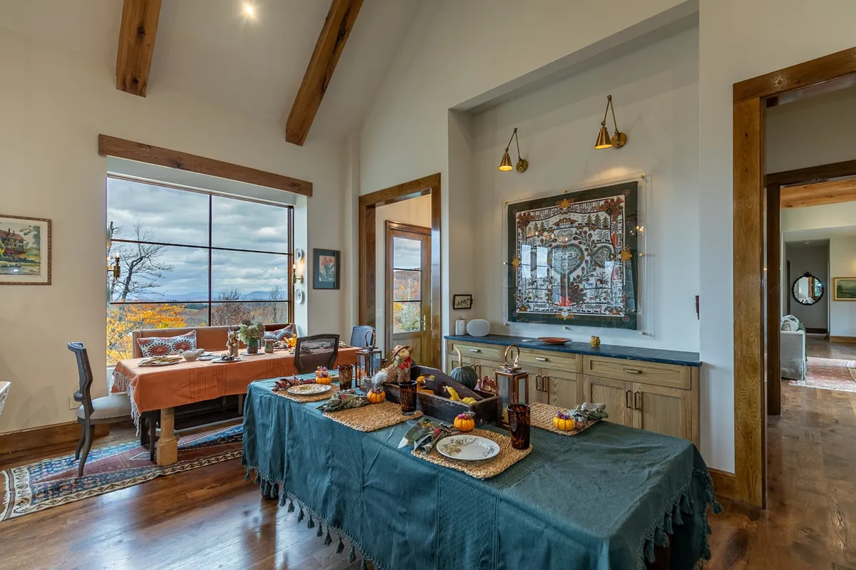 A dining table set with plates and decorations sits by a large window offering a scenic view, complemented by wall artwork. The setup suggests a space ready for family gatherings or festive meals, with the outdoors always in view.