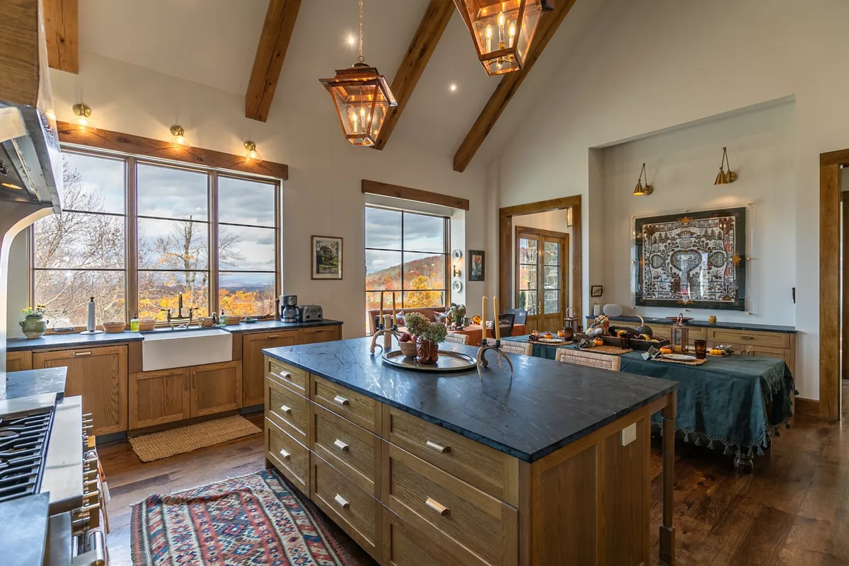 A spacious kitchen with a large kitchen island, panoramic window view, wooden beams, and decorative lanterns. The abundance of natural light and scenic vistas create a seamless connection to the outdoors, hinting at the transition between interior cooking and exterior dining experiences.