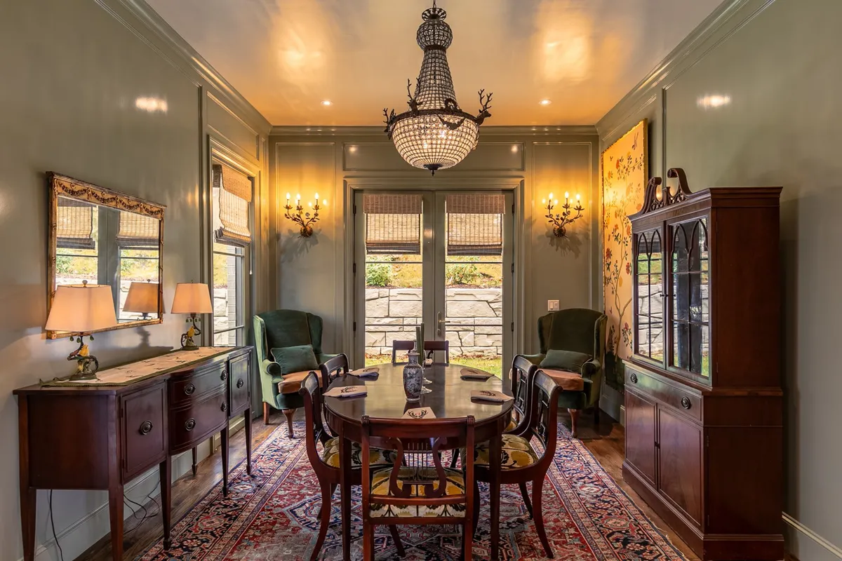 A spacious, elegant dining room featuring a large table set beneath a sophisticated chandelier. Ample windows flood the space with natural light, and tasteful wall art and a buffet table create an inviting environment for group gatherings or celebratory meals.