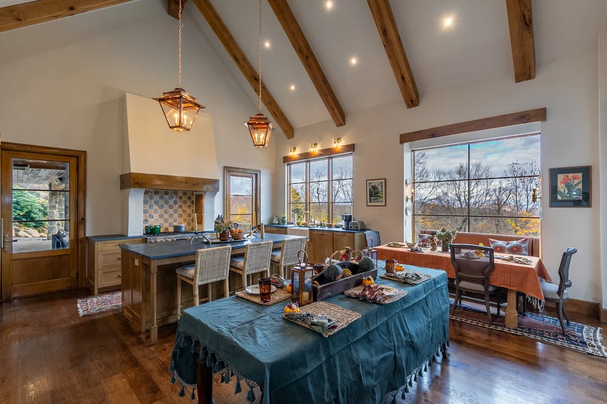 A warm, inviting dining area centered around a large table beneath stylish light fixtures and exposed wooden beams. Wide windows offer beautiful mountain views, and the setup is ideal for large family meals, festive celebrations, or casual morning coffee.