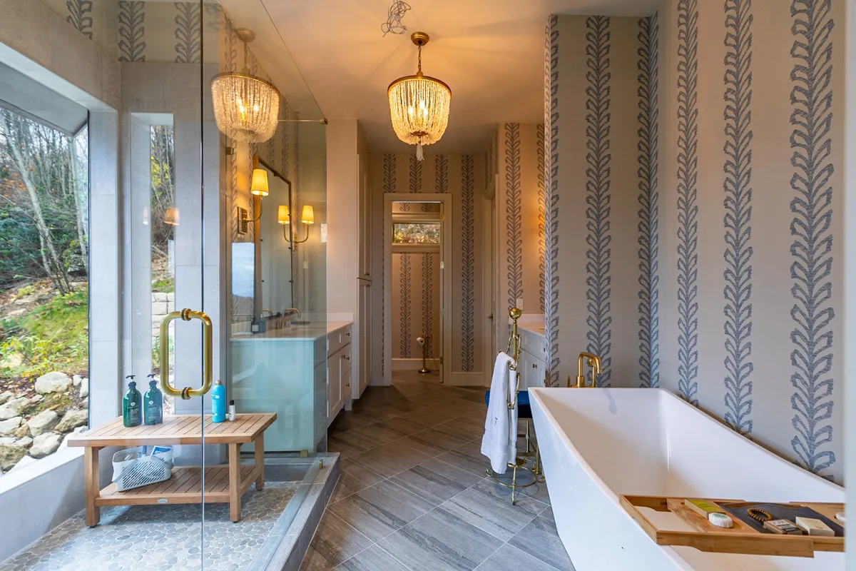 An elegant, spa-like bathroom featuring a freestanding bathtub, large glass shower enclosure, chandeliers, and decorative wallpaper. Natural light fills the space, enhancing its tranquil and luxurious atmosphere.