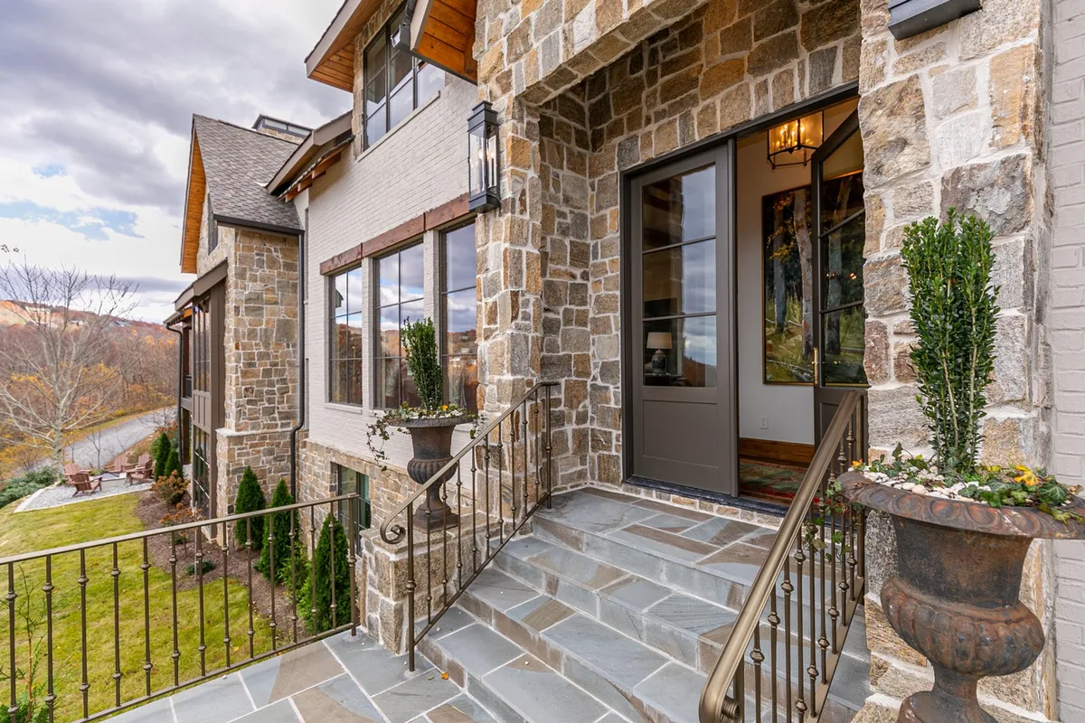 A warm and inviting exterior view of a stone-faced entrance, flanked by planters and illuminated by lanterns. The stairs lead up to a cozy porch, hinting at a welcoming home environment. The soft lighting suggests a comfortable ambiance, perfect for returning from a day of exploring the nearby mountain towns.